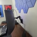 A home office setup with a curved monitor on an adjustable arm, keyboard, and blue geometric acoustic panels on the wall