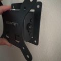 A black wall-mounted bracket or holder with "MOUNT IT!" text visible, being held by a person's hand against a white wall. The bracket features multiple mounting holes and appears to be designed for securing devices or equipment.