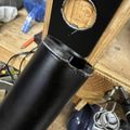 A black cylindrical container or pipe positioned vertically in a wooden workshop setting with tools and equipment visible in the background