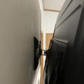A TV mounted on a white wall with visible mounting bracket and cables in what appears to be a narrow hallway or corridor