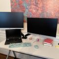 A modern home office setup with two monitors, a laptop, wireless keyboard and mouse on a white desk, with colorful abstract artwork on the wall behind