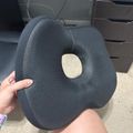 A person holding a dark gray ergonomic seat cushion with a circular cutout in the center, designed for comfort while sitting