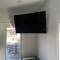 A flat screen TV mounted on a white wall in a corner room with a window showing outdoor patio furniture and some items on a shelf below the TV