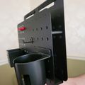 A black perforated metal organizer or holder with multiple compartments and cup holders, being held by a hand against a white wall background