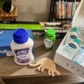 A table with various household items including Lysol disinfectant wipes, laundry sanitizer, Centrum vitamins, Cetaphil skincare products, and a wooden toy dinosaur, with bookshelves and a TV wall mount box visible in the background