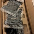 Cardboard shipping box containing multiple plastic-wrapped packages of dark-colored clothing or textiles stacked inside.