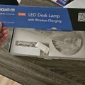 An opened box containing a Mount-It LED desk lamp with wireless charging capability, showing the lamp components and cables inside the packaging on a wooden surface