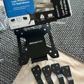 A MOUNT IT low-profile tilt TV wall mount package displayed on a gray textured fabric surface, showing the black metal mounting bracket, instruction manual with blue packaging, and four black wall anchor brackets with handles.