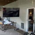 Covered outdoor patio with mounted TV, wooden table, and glass doors leading to an indoor office space