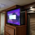 Interior of an RV showing a flat screen TV mounted in a wooden entertainment center with purple display screen, wooden cabinetry, and window blinds visible