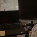 A laptop computer sits on a desk with papers, a coffee mug, and office supplies, with a whiteboard visible on the wall in the background