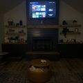 A modern living room with a wall-mounted TV displaying a streaming service menu, flanked by wooden shelves holding decorative items and plants. A fireplace is centered below the TV, with a round ottoman containing blankets and snacks in the foreground on a patterned area rug.