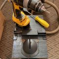 A yellow power drill mounted on a metal drilling jig or guide positioned on a carpeted floor with a vacuum hose nearby