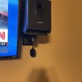 A black Logitech device mounted on a wall bracket next to a TV screen displaying news content