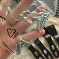 A person's hand holding an open plastic package containing metal screws and black hardware brackets with oval cutouts, displayed on a light-colored textured surface.