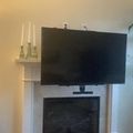 Living room with flat screen TV mounted above white fireplace, decorative candles on mantel shelf, and houseplant visible on right side