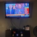 A television mounted on a wall displaying a news program with two men in suits, with a media console below and a fan visible to the right