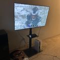 A large TV mounted on a black stand displaying a football game, with a gaming console below and a small dog lying on the carpet in front