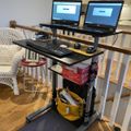 Standing desk setup with two laptops, keyboard, and mouse positioned over a treadmill in a home office space with white railings and kitchen visible in background