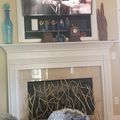 Living room with white fireplace mantel, mounted TV displaying a show, decorative items on shelves, and a dog resting on a cushion in front of the fireplace