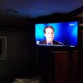 A television screen displaying a news broadcast with a male anchor against a blue background, mounted on a wall in a dimly lit room with wooden furniture and blinds visible.