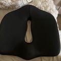 Black ergonomic seat cushion with cutout center placed on a bed with decorative pillows