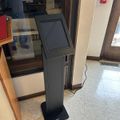 Black digital kiosk or information terminal standing on tiled floor in what appears to be a public building or office lobby