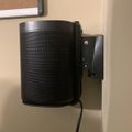 Black cylindrical smart speaker mounted on wall with metal bracket and power cord