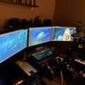 A computer workstation with three monitors displaying underwater ocean scenes, surrounded by audio equipment including keyboards and speakers in a dimly lit room