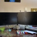 A dual monitor computer setup on a desk with various office supplies including colorful markers, papers, and a green mug, with artwork visible on the wall behind