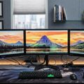 A modern office workspace with three curved monitors displaying scenic mountain landscapes, positioned on a desk with keyboard, mouse, and headphones, with diplomas and certificates hanging on the wall behind