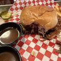 A sandwich with meat and onions on a bun served on red and white checkered paper with a small cup of au jus and pickle slices