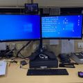 A dual monitor computer workstation setup on a desk with keyboard, mouse, papers, and office equipment in what appears to be a workplace environment