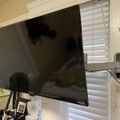 A flat screen TV mounted on a wall arm in a room with white blinds and various items on shelves