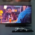 A large flat-screen TV mounted on a wall displaying a scene with a person sitting at a table, with a decorative shelf below containing what appears to be a metallic sculpture