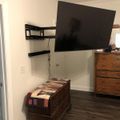 A bedroom corner with a wall-mounted TV, wooden chest with patchwork quilt, and tall wooden dresser on hardwood flooring
