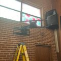 A wall-mounted TV and speaker system installed on a brick wall with a yellow ladder positioned below during setup or maintenance work