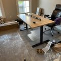 A home office setup with a wooden standing desk, black office chair, and assembly materials scattered on gray carpet near a window