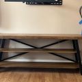 A wooden TV stand with black metal frame and cross-bracing, featuring two open shelves, positioned against a beige wall with a TV mount visible above