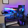 A modern gaming setup with dual monitors displaying colorful space imagery, RGB lighting, gaming chair, desk with keyboard and microphone, and a framed certificate on the wall
