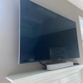 A flat-screen television mounted on a white wall above a decorative white mantelpiece, with a white soundbar and black cables visible beneath it.