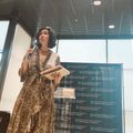 Woman in patterned dress speaking into microphone at podium with educational institution backdrop