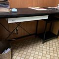 A black desk with papers and supplies on top, positioned against a wood-paneled wall with a patterned floor underneath