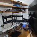 A home entertainment setup with wooden shelves holding various electronic devices, cables, and equipment beneath a wall-mounted TV