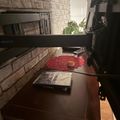 A dark wooden desk with a laptop, red decorative mat, and small cup, positioned against a brick wall near a fireplace with a TV mounted above