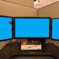 A modern workspace with three large computer monitors displaying blue screens arranged on a curved black desk, with a white device and keyboard visible on the desktop surface.