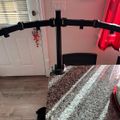 A black curved monitor stand or mount positioned in a home interior with white doors, wooden flooring, a patterned rug, and red curtains visible in the background.