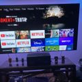 A smart TV displaying a streaming service interface with various app icons including YouTube, Netflix, Disney+, Prime Video, and HBO Max, set up in a modern living room with purple ambient lighting