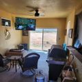 Interior view of a living room with a ceiling fan, mounted TV showing sports, sliding glass door overlooking an open field, furniture including chairs and a grill, with peeling paint visible on the ceiling