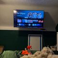 A smart TV mounted on a wall displaying a streaming service menu with "The Paper" show featured prominently. Below the TV is a bed with green bedding and a tan-colored blanket, along with cleaning supplies including an orange spray bottle and cloth. The room has textured walls and various items on shelves to the right.